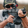 Person holding a rifle with a red dot sight, wearing tactical gear.