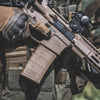 Close-up of a person holding a rifle with a magazine, wearing tactical gloves and gear.