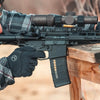 Person holding a rifle with a scope, wearing gloves, on a wooden surface.